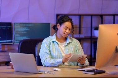 Genç Afro-Amerikalı soru cevap mühendisi geceleri ofiste tablet bilgisayarla çalışıyor.