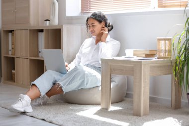 Genç Afro-Amerikalı soru cevap mühendisi dizüstü bilgisayarlı evde armut koltukta oturuyor.