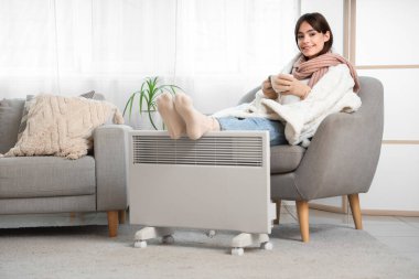 Evdeki elektrikli konteyner ısıtıcısında ayaklarını ısıtan koltukta çay içen genç bir kadın.