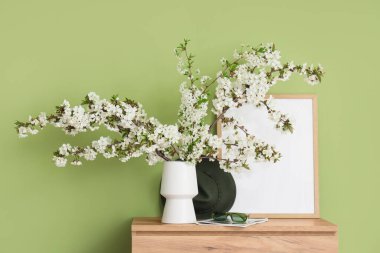 Çiçek açan dalları, güneş gözlükleri ve yeşil duvarın yanındaki çekmecelerin üzerindeki çerçeveli vazo.
