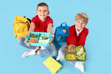 Beslenme çantaları ve sırt çantalarıyla mavi arka planda oturan küçük okul çocukları.