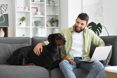 Dobermann köpeği ve dizüstü bilgisayarı olan genç adam evdeki kanepede oturuyor.