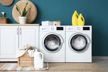 Çamaşır makineleri, deterjan şişeleri ve hasır sepeti olan modern çamaşır odası.