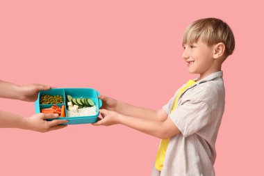 Küçük çocuk pembe arka planda kadın ellerinden beslenme çantası alıyor.