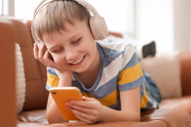 Modern cep telefonu ve kulaklıkları olan sevimli küçük çocuk evdeki kanepede müzik dinliyor.