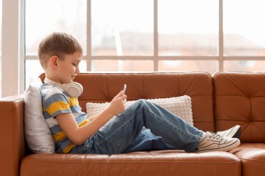 Modern tablet bilgisayarı ve kulaklıkları olan sevimli küçük çocuk evdeki kanepede yatıyor.