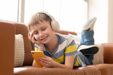 Modern cep telefonu ve kulaklıkları olan sevimli küçük çocuk evdeki kanepede müzik dinliyor.