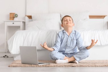 Modern dizüstü bilgisayarı olan şirin bir çocuk yatak odasında meditasyon yapıyor.