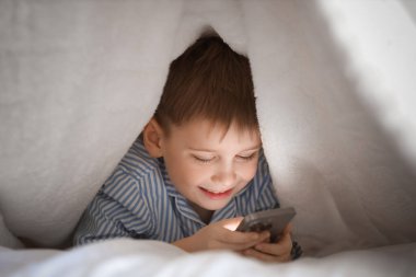 Küçük şirin çocuk battaniyenin altında modern cep telefonu kullanıyor.