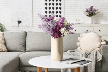 Oturma odasındaki masa üzerinde leylak çiçekleri ve dergiler olan vazo.