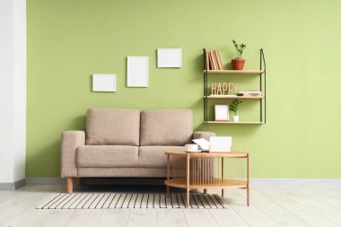 Oturma odasının iç tarafındaki yeşil duvarın yanındaki tahta sehpa ve gri kanepe.