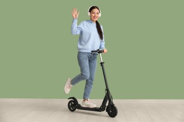 Kulaklıklı güzel bir Asyalı kadın yeşil duvarın yanında modern elektrikli scooter kullanıyor.