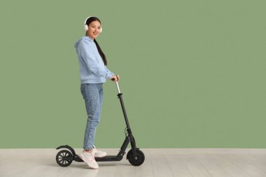 Yeşil duvarın yanında modern elektrikli scooterlı kulaklıklı güzel bir Asyalı kadın.