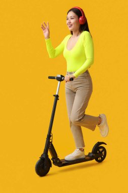 Sarı arka planda modern elektrikli scooter kullanan kulaklıklı güzel bir Asyalı kadın.