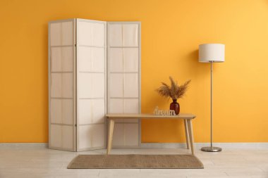 Sarı duvarın yanında katlanır ekran, bank ve zemin lambası olan odanın içi.