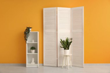 Odanın içinde katlanan ekran, raf ünitesi ve sarı duvarın yanındaki ev bitkileri.