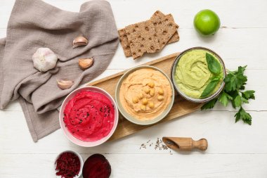 Beyaz ahşap arka planda sarımsak, maydanoz, pancar ve kraker içeren farklı lezzetli humus kaseleri.