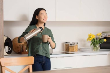 Asyalı kadın mutfakta termostan fincana sıcak çay dolduruyor.
