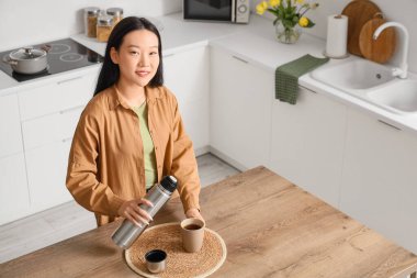 Asyalı kadın mutfakta termostan fincana sıcak çay dolduruyor.