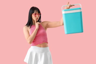 Pembe arka planda taşınabilir buzdolabı olan düşünceli genç bir kadın.