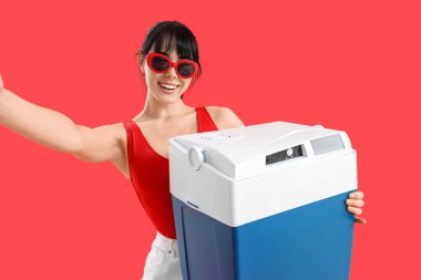 Portatif buzdolabı taşıyan genç bir kadın kırmızı arka planda selfie çekiyor.