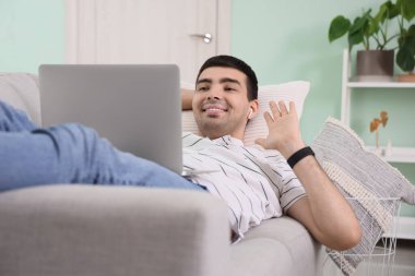 Dizüstü bilgisayarlı genç adam evdeki kanepede sohbet ediyor.