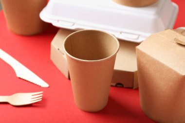 Paket kâğıt bardak, kaplar ve çatal bıçak takımı kırmızı arka planda, yakın plan.