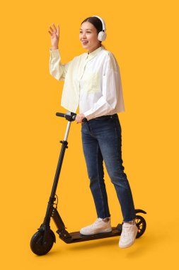 Sarı arka planda modern elektrikli scooterlı kulaklıklı güzel bir Asyalı kadın.