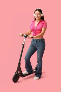 Pembe arka planda modern elektrikli scooterlı kulaklıklı güzel bir Asyalı kadın.