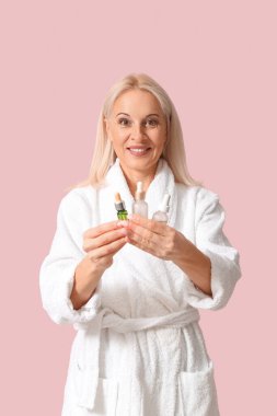 Bornozlu olgun bir kadın pembe arka planda kozmetik yağı şişeleri tutuyor.