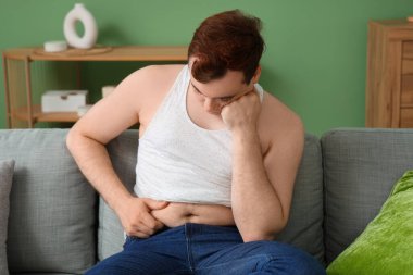 Dar kot pantolonlu, evdeki kanepede oturan üzgün, şişman bir adam. Kilo alma kavramı