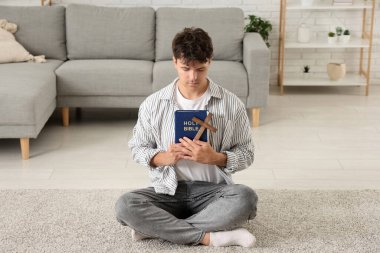 Ahşap haçlı genç adam ve kutsal kitap oturma odasında dua ediyor.