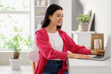 Evdeki masada dizüstü bilgisayar kullanan genç bir kadın.