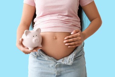 Mavi arka planda domuz kumbarası olan genç hamile kadın, yakın plan. Annelik Faydası konsepti