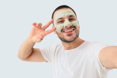 Yüzünde kozmetik balon maskesi olan yakışıklı genç adam gri arka planda selfie çekiyor.