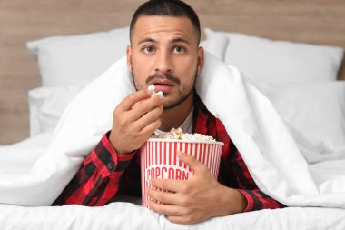 Patlamış mısırlı genç adam yatakta film izliyor.