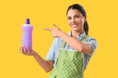 Güzel ev hanımı sarı arka plandaki deterjan şişesini işaret ediyor.