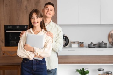 Dizüstü bilgisayarlı genç çift mutfakta sarılıyor.