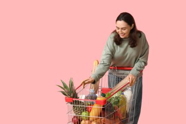 Pembe arka planda alışveriş arabası olan güzel, genç ve mutlu bir kadın.