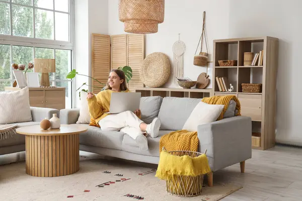 Aydınlık oturma odasındaki gri kanepede dizüstü bilgisayarı olan güzel genç bir kadın.