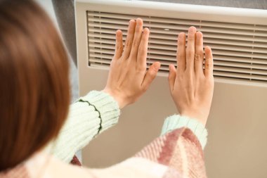 Evde elektrikli konteyner ısıtıcısının yanında ellerini ısıtan genç bir kadın var.