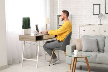 Genç adam iş yerinde oturup evde dizüstü bilgisayarla çalışıyor.