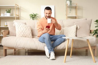 Oturma odasında cep telefonu kullanarak çay içen yakışıklı genç adam.