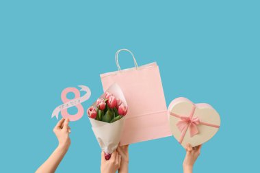 Elinde kartpostal, kağıt poşet ve mavi arka planda hediye kutusu olan çiçekleri tutan kadın eller. Uluslararası Kadınlar Günü için Alışveriş