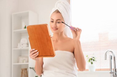 Havluya sarılı güzel bir kadın banyoda aynaya bakarken maskara sürüyor.