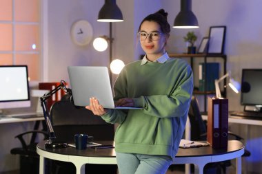 Akşam ofiste dizüstü bilgisayarla çalışan genç bir kadın.