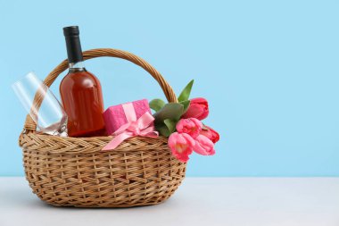 Laleli sepet, şarap şişesi ve Anneler Günü için hediye paketi mavi arka planda beyaz masada.