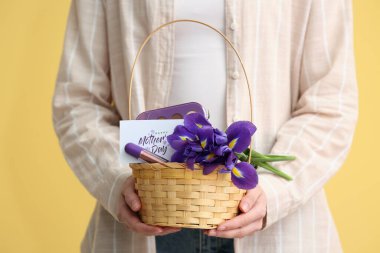 Sarı arkaplanda anneler günü için çiçek, kartpostal ve kozmetik taşıyan sepet taşıyan genç bir kadın.