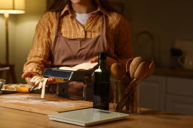 Genç bir kadın karanlık mutfakta makineyle makarna yapıyor.