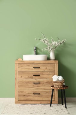 Banyonun içinde, yeşil duvarın yanındaki çekmecelerin üzerinde çiçek açan bir vazo var.